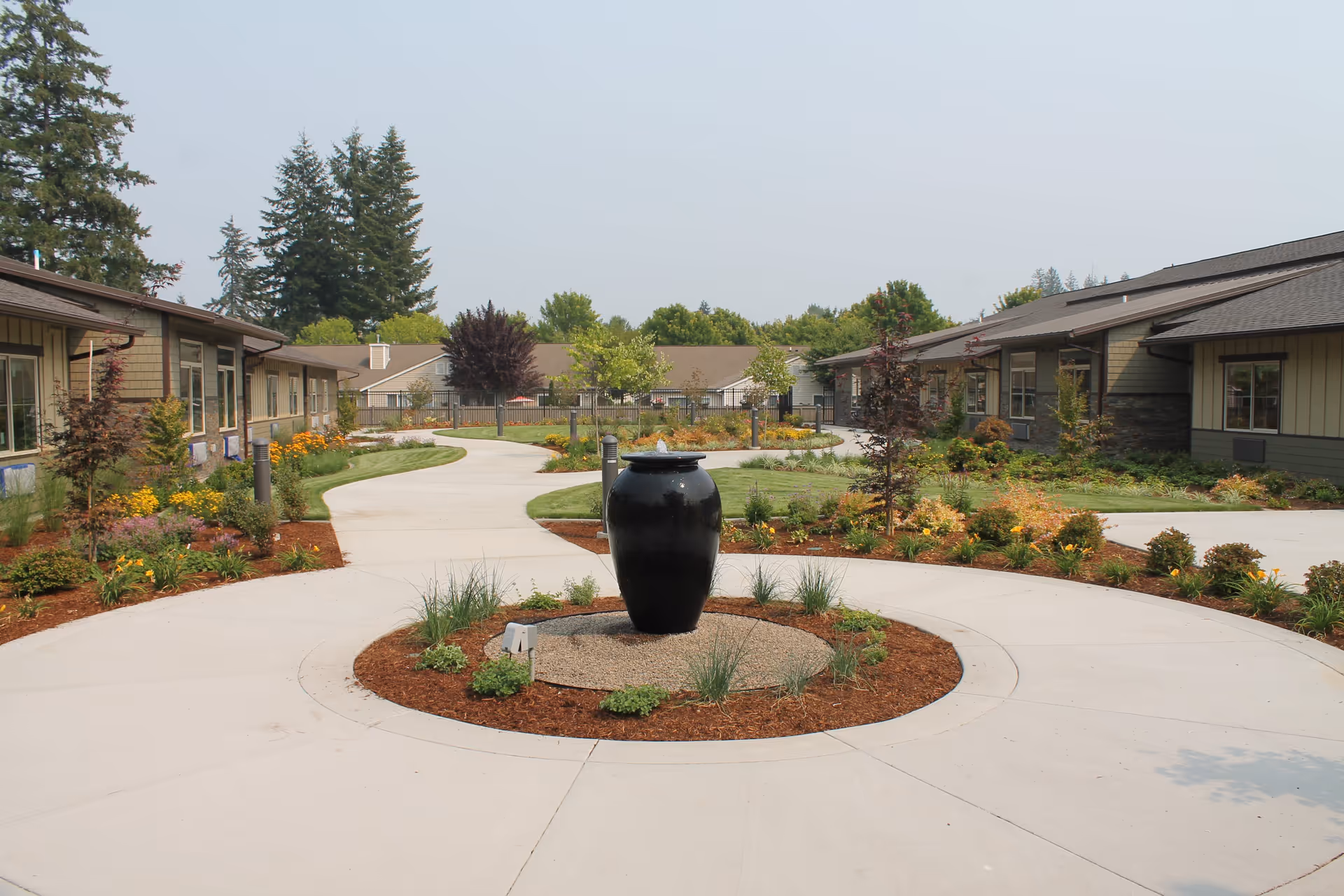 Outdoor courtyard area at Fieldstone Memory Care Olympia featuring a circular concrete pathway with a large black urn in the center surrounded by plants and mulch. The courtyard is landscaped with various shrubs, flowers, and small trees, and is bordered by single-story buildings with windows. Tall evergreen trees are visible in the background under a clear sky.