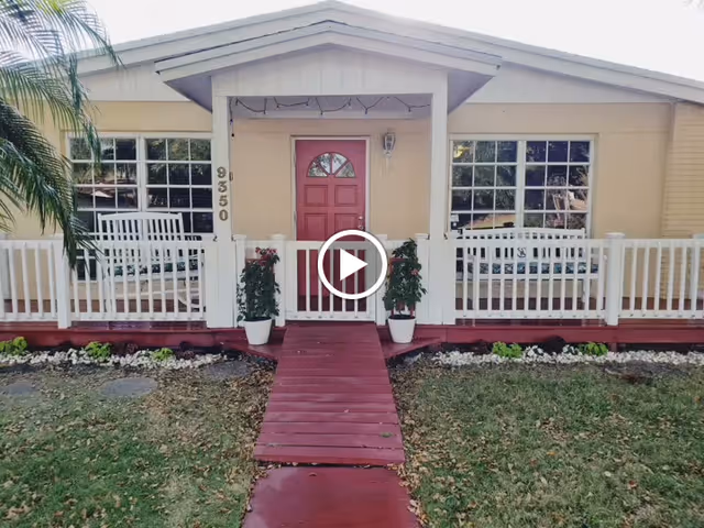 Front exterior view of a single-story house with a red door, white railing, two potted plants on either side of the entrance, and a red wooden ramp leading to the door. The house number 9350 is visible on a post near the door. There are two large windows on either side of the door and a small garden with grass and plants in front.