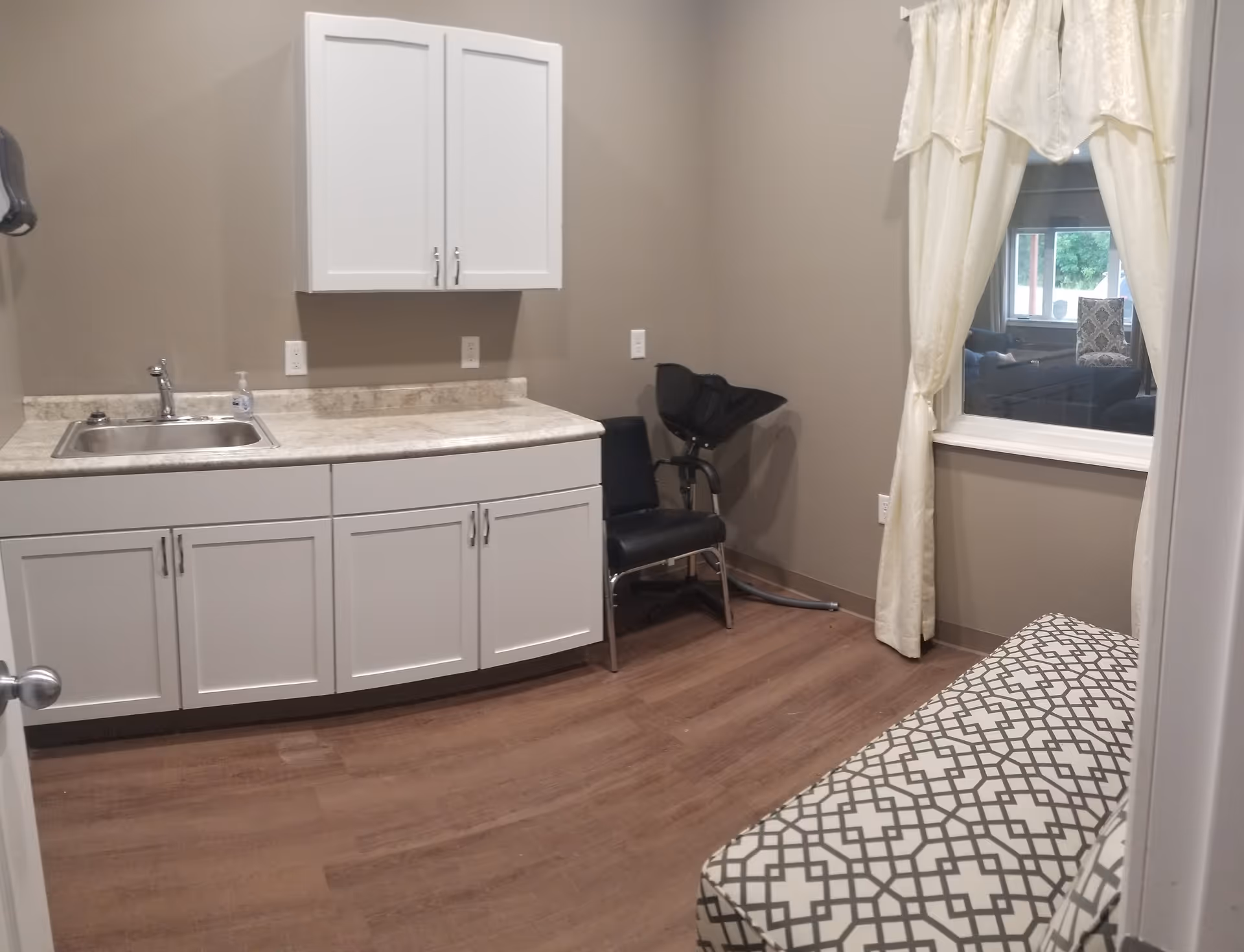 Small interior room with white cabinets and sink, a patterned bench, a black salon chair, and a curtained window.