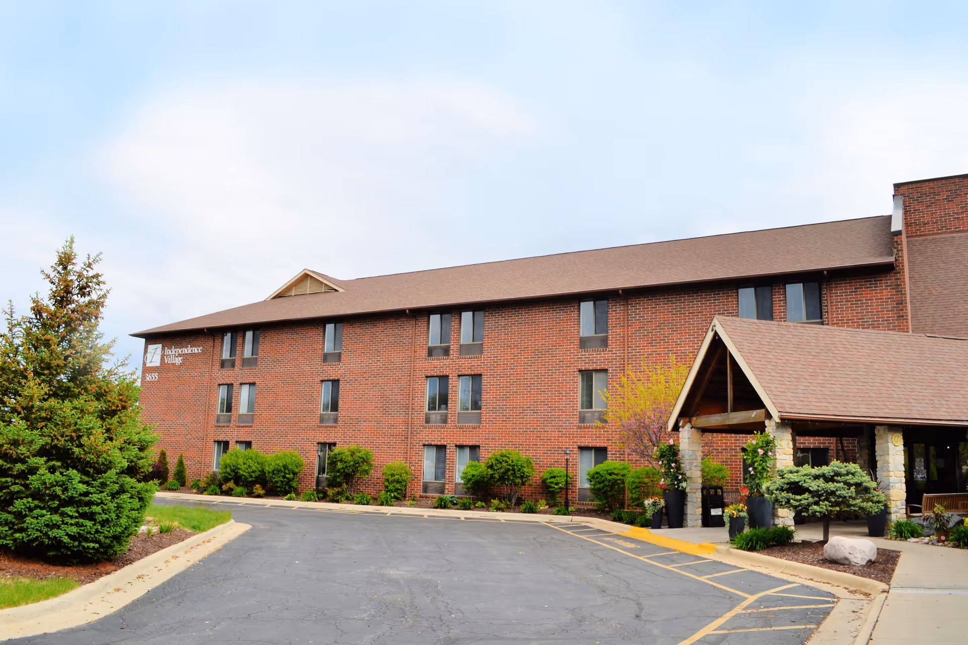 Exterior view of a three-story brick building with multiple windows and a covered entrance supported by stone pillars. The building is surrounded by landscaped greenery including bushes and trees, with a paved driveway and parking area in front. The building has a sign that reads 'Independence Village' along with the number 3655.