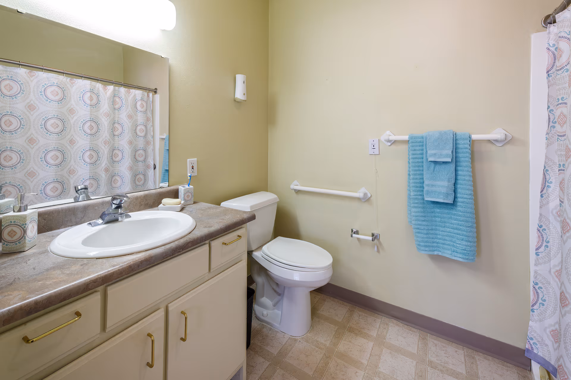 A clean bathroom with a white sink and countertop, a toilet, a large mirror above the sink, a shower curtain with a circular pattern, and blue towels hanging on a towel rack.