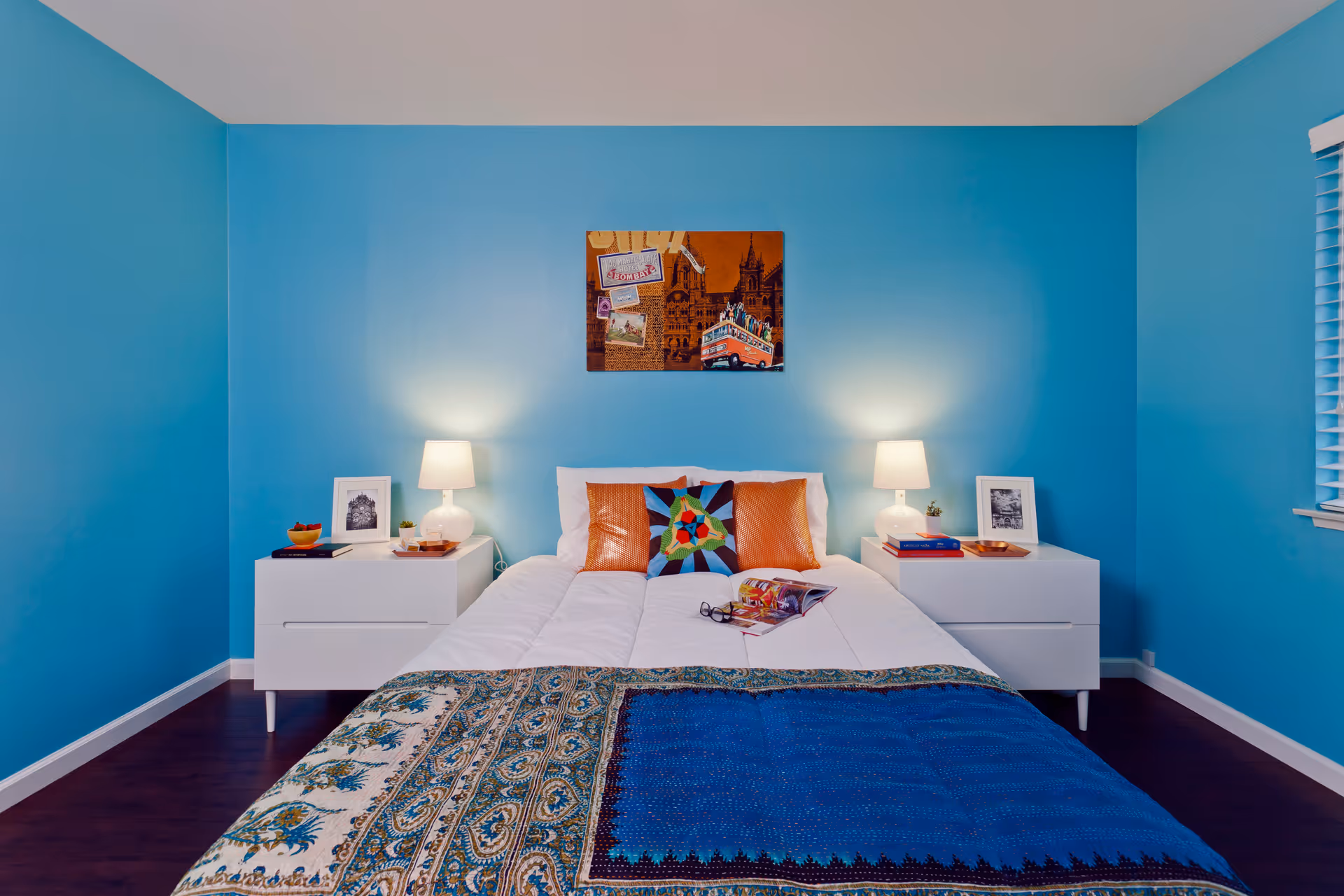 A neatly made bed with white bedding and decorative pillows is centered against a bright blue wall. On either side of the bed are white nightstands, each with a lamp, framed pictures, and small decorative items. Above the bed hangs a colorful piece of artwork. The room has dark wood flooring and a window with blinds on the right side.