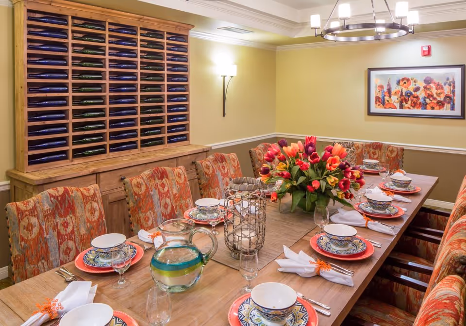 A dining room with a long wooden table set for eight people. The table is decorated with colorful plates, bowls, glasses, white napkins with orange napkin rings, a glass pitcher with water, two candle holders, and a vibrant bouquet of tulips in the center. The chairs have patterned upholstery in shades of orange and red. On the wall behind the table is a large wooden wine rack filled with bottles and a framed floral painting. The room is softly lit by a chandelier and wall sconces.
