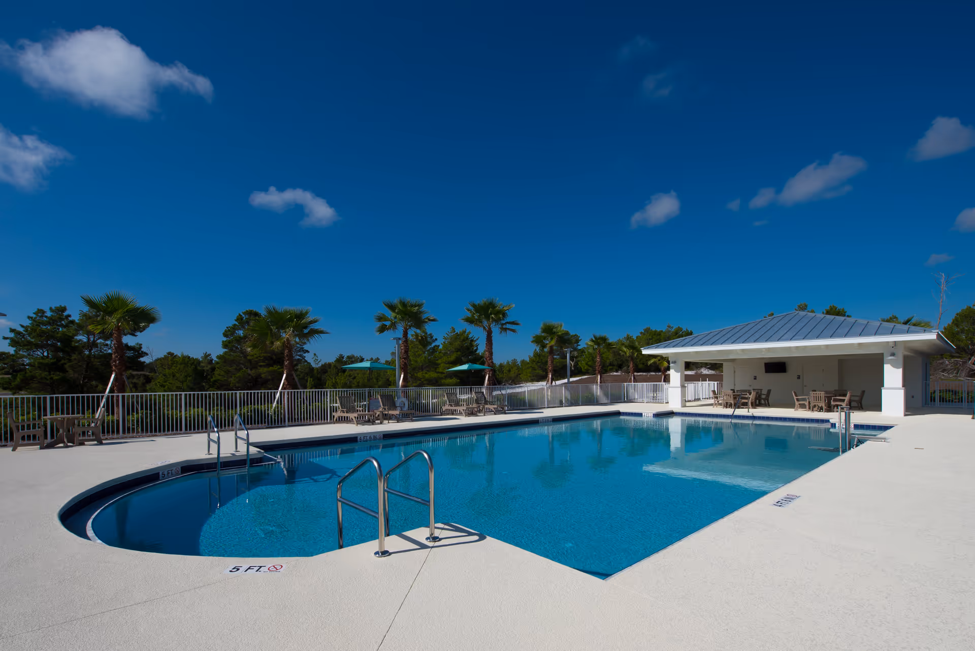 Outdoor swimming pool with deck chairs, a covered pavilion, and palm trees under a bright blue sky.
