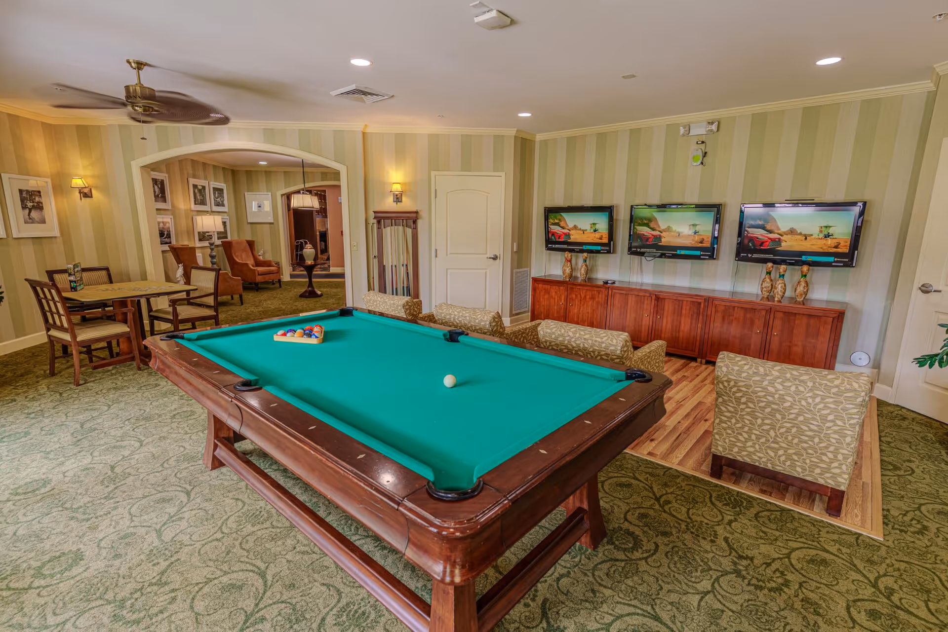 A recreation room featuring a pool table in the foreground, seating area, three wall-mounted TVs and a small dining table.