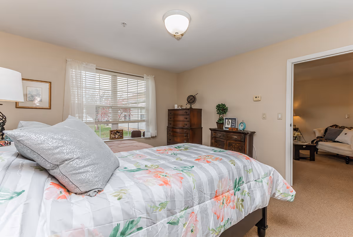 A bedroom with a bed covered in a floral patterned comforter and silver pillows. There is a window with white blinds and sheer curtains letting in natural light. Two wooden dressers are against the wall, one with decorative items including a globe and framed photos. A doorway leads to a living room area with a beige couch and a lamp.