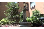 Outdoor courtyard featuring a stone pedestal fountain surrounded by hostas and shrubs beside a brick building.