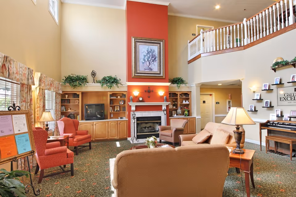 A cozy living room area with high ceilings, featuring a fireplace with a red accent wall and a framed floral painting above it. The room has built-in wooden shelves on either side of the fireplace, decorated with plants and various items. There are several upholstered chairs and sofas arranged around a central coffee table with a flower arrangement. A lamp on a side table provides warm lighting. Large windows with floral curtains allow natural light to enter. On the right side, there is a wall with small shelves displaying framed photos and a sign that reads 'Gallery of Excellence'.
