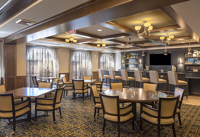 A spacious dining area in a senior living facility featuring multiple round and square tables with cushioned chairs. The room has large windows with sheer curtains allowing natural light to enter. There is a bar counter with high chairs and a large flat-screen TV mounted on the wall behind it. The ceiling has recessed lighting and decorative chandeliers, and the floor is covered with a patterned carpet.