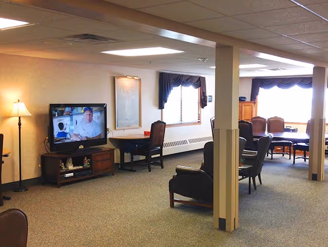 A spacious common area in a senior living facility with multiple seating options including armchairs and dining chairs around tables. A television on a wooden stand is on, showing a man and a child. The room has beige walls, carpeted floor, and windows with dark valances letting in natural light. A floor lamp is also visible near the TV.
