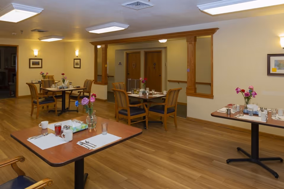 Dining room with set tables and chairs, wood flooring, and vases of flowers in an assisted living facility.