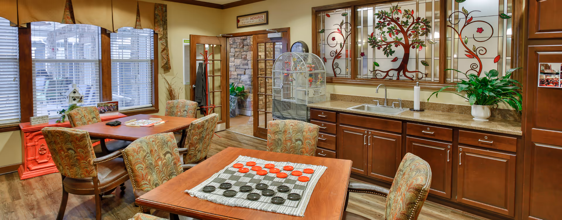 Cozy senior living common room with wooden tables and upholstered chairs, a kitchenette with stained-glass window, and a large birdcage.