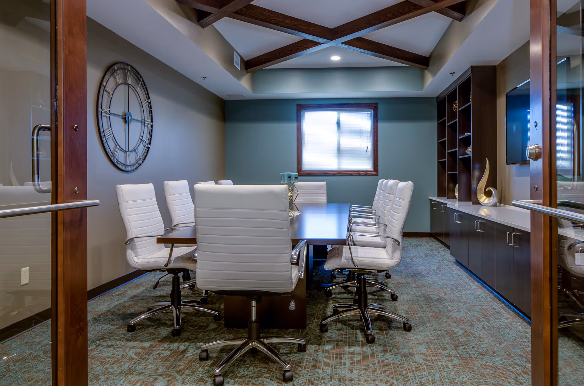 Conference room with a long table surrounded by white swivel chairs, a wall clock, shelving, and a window.