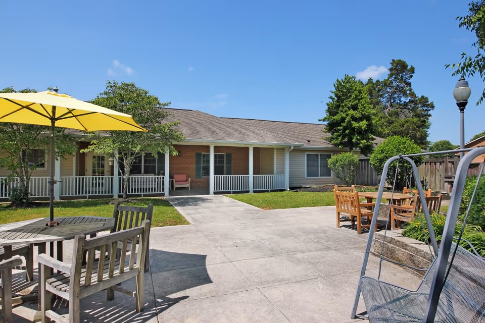 Sunny outdoor courtyard with patio tables and chairs, a yellow umbrella and a swing in front of a single-story building with a covered porch.