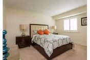 A neatly made bed with a patterned gray and white bedspread and three orange accent pillows in a bright bedroom. The bed has a padded headboard and is flanked by two dark wooden nightstands, each with a lamp. A window with white blinds and a framed picture on the wall are visible.