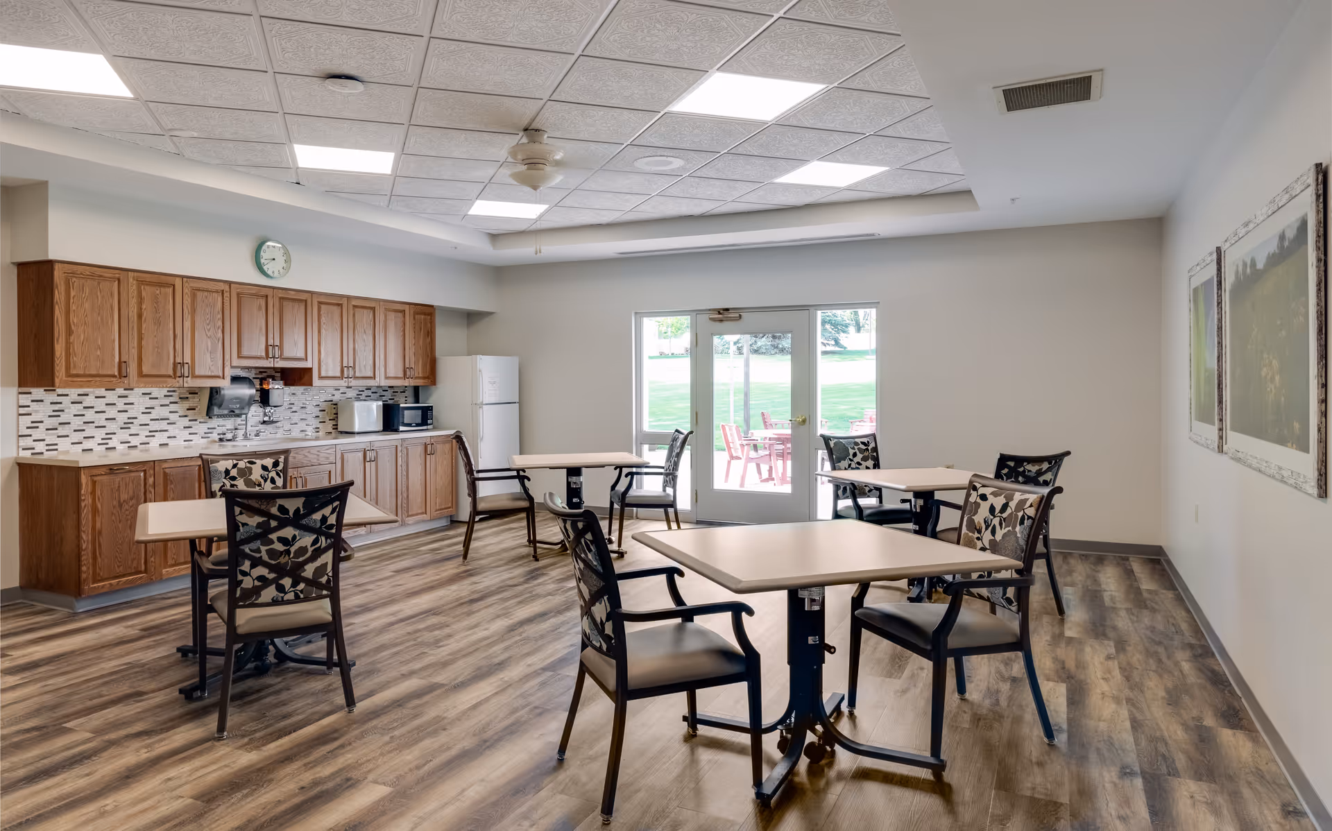 Bright communal dining area with a kitchenette, several tables and chairs, and glass doors opening to an outdoor patio.