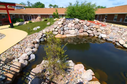 Outdoor courtyard area at Addington Place of Des Moines featuring a pond surrounded by rocks and a small waterfall. The courtyard is bordered by a single-story building with a red roof and windows, and there is a paved walkway with a red pergola structure on the left side.