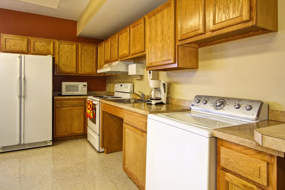 A kitchen area with wooden cabinets, a white refrigerator, microwave, stove with oven, coffee maker, and a washing machine. The walls are painted beige and one wall is a dark reddish-brown color.
