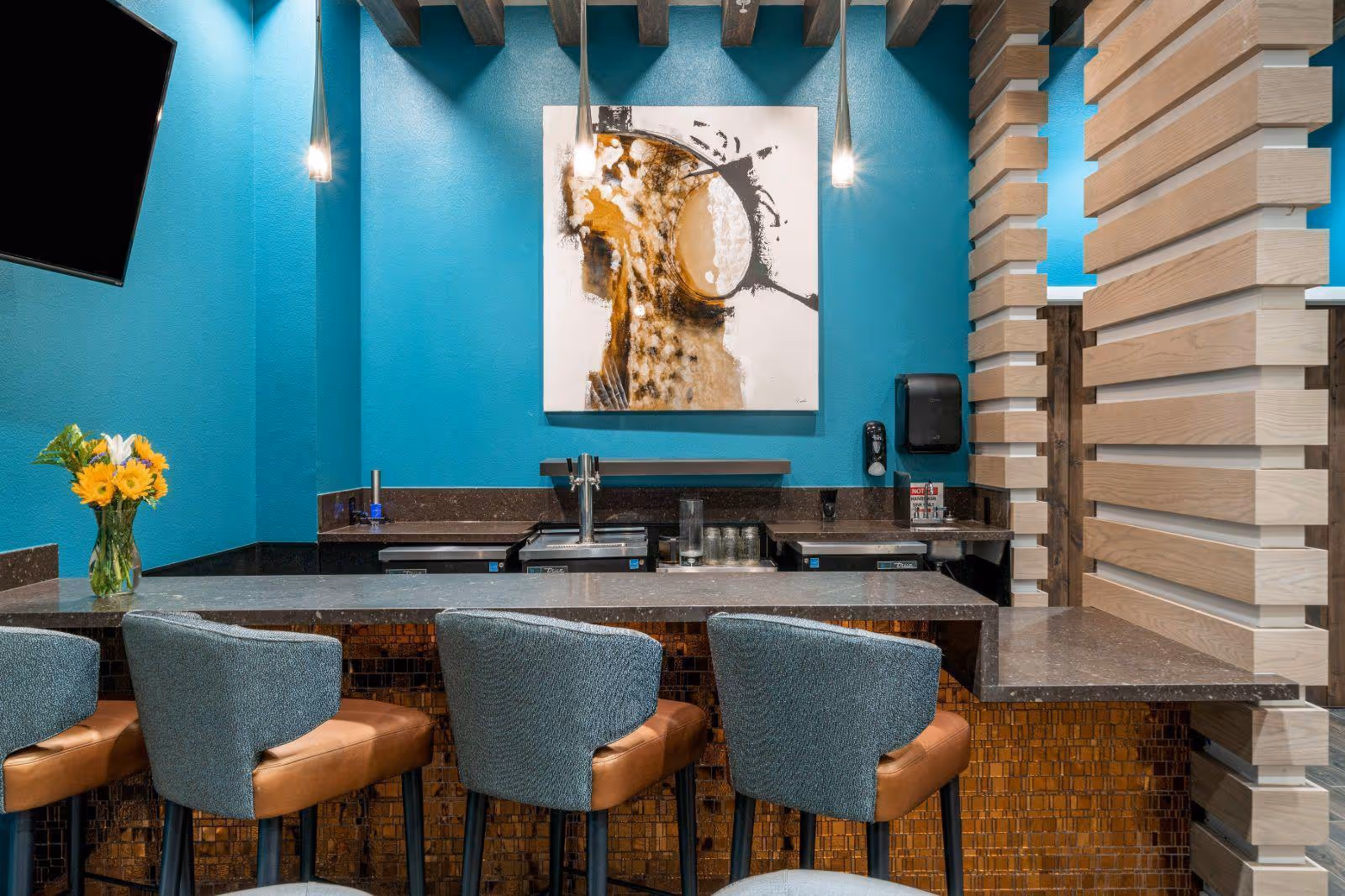 A modern indoor bar area with teal walls, a countertop with four upholstered stools, pendant lights, artwork, and a vase of yellow flowers.