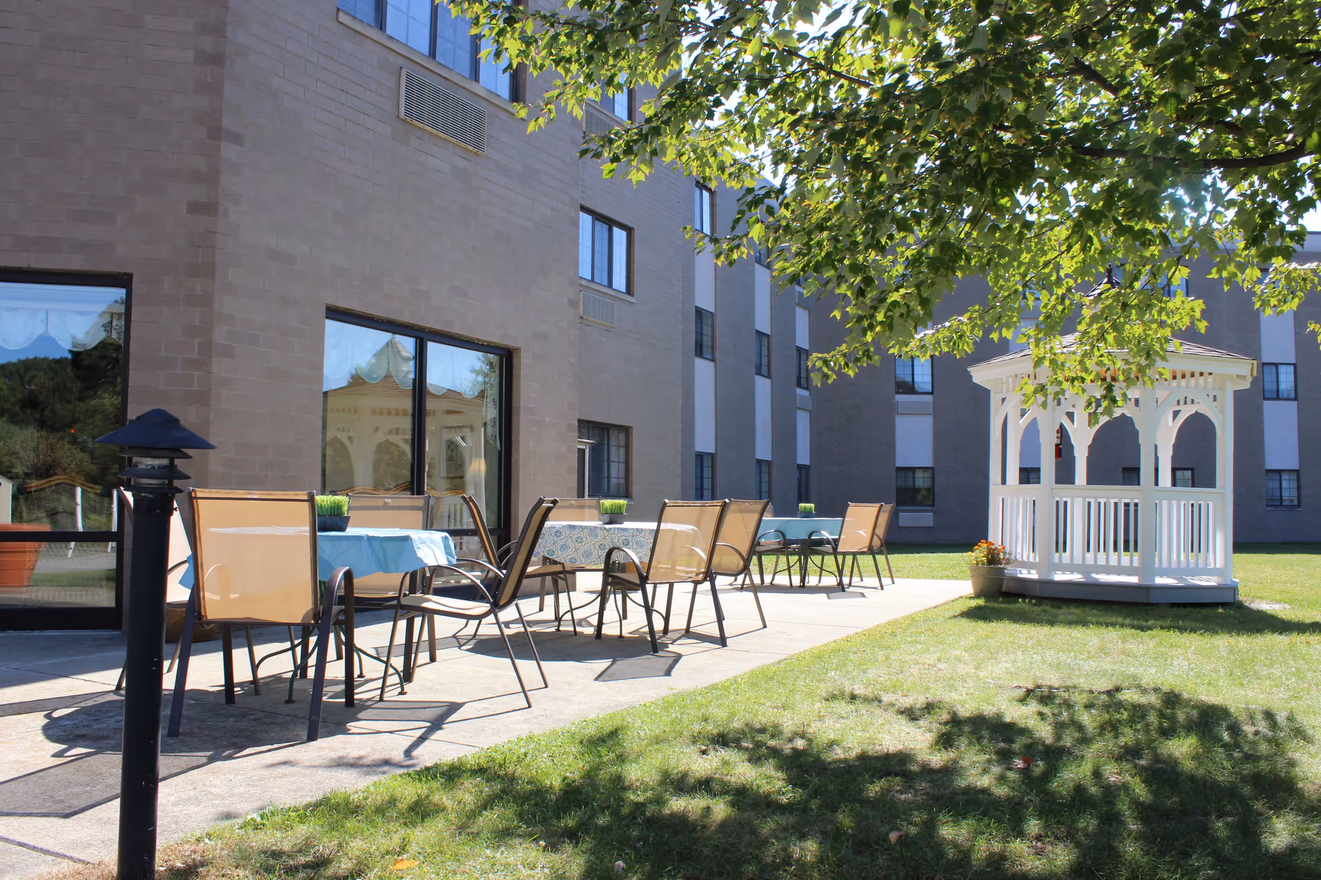 Outdoor patio area at The Residence at Arrowood featuring several tables with chairs on a concrete walkway next to a building. A white gazebo is visible on the grassy lawn, and tree branches provide partial shade.