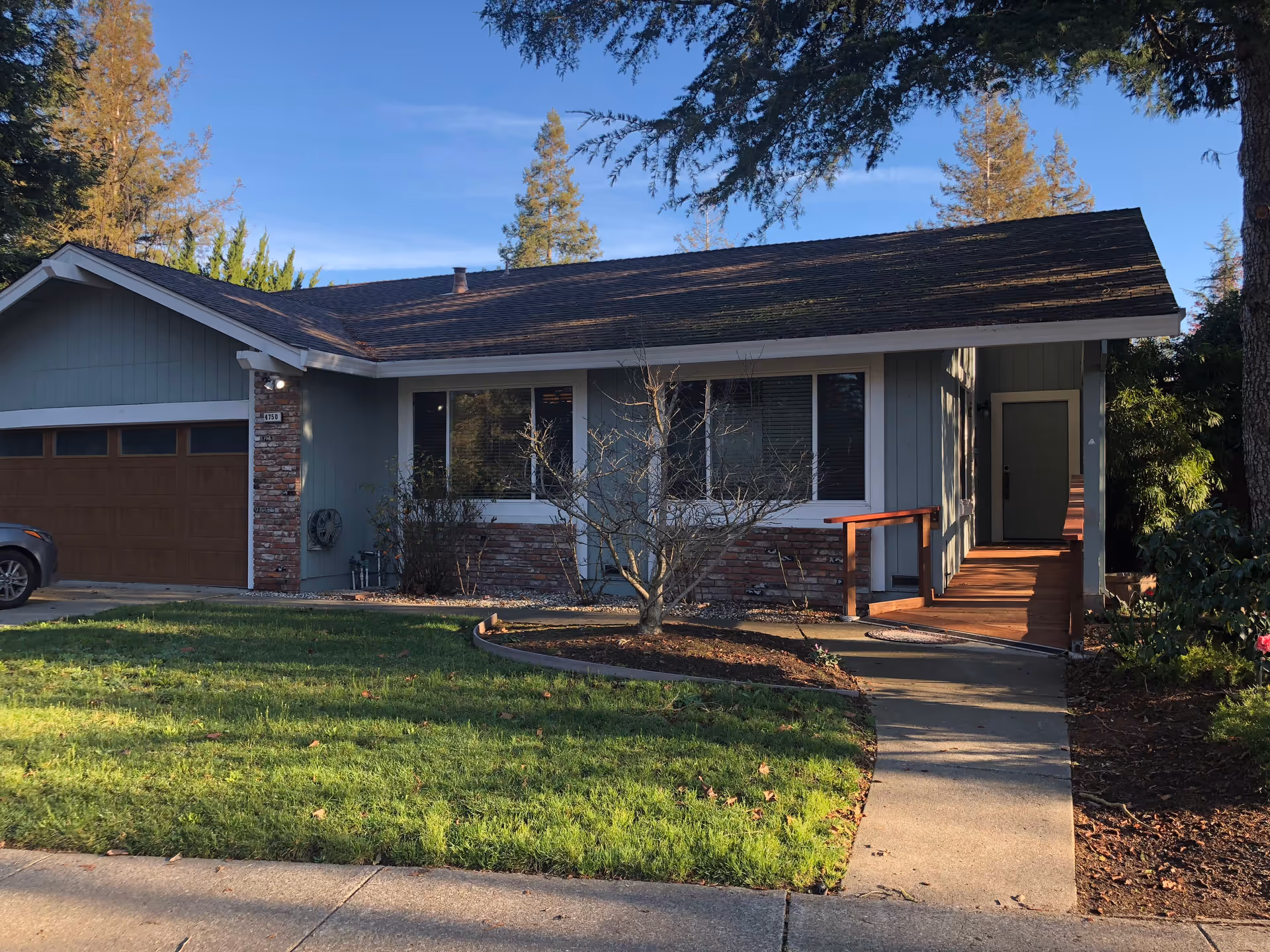 Front exterior view of a single-story house with a wooden garage door, large windows, a small porch with a ramp, and a well-maintained lawn with trees and shrubs under a clear blue sky.