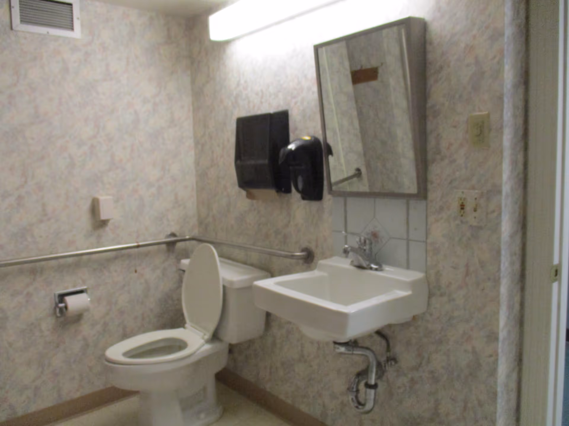 Small institutional bathroom with a toilet, wall-mounted sink, mirror, grab bars, and paper dispensers.