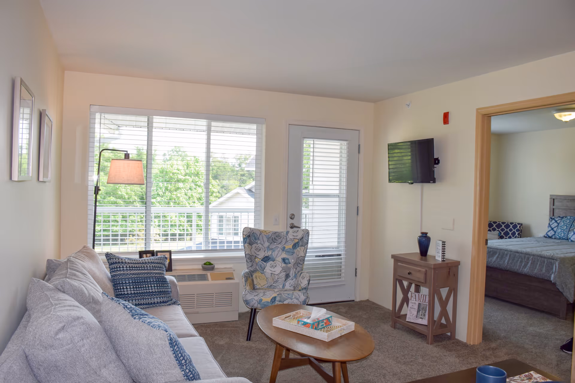A cozy living room with a light gray sofa adorned with blue patterned pillows, a floral armchair, a wooden coffee table with a tissue box, and a small wooden side table with a blue vase and decorative items. A flat-screen TV is mounted on the wall. Large windows with blinds and a glass door let in natural light and show greenery outside. An adjoining bedroom with a bed and blue pillows is visible through an open doorway.