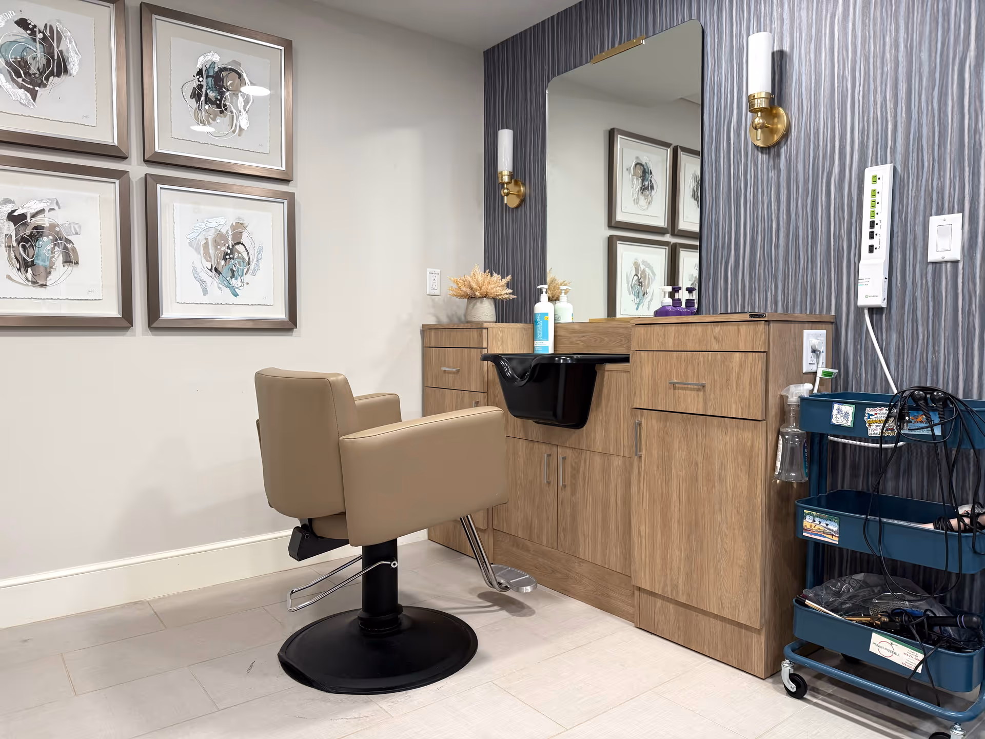 A salon styling station with a tan swivel chair in front of a wooden vanity, mirror, sink and a rolling cart with hair tools.