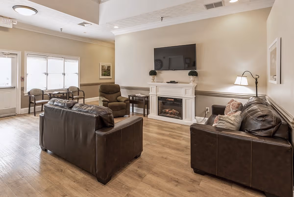 A cozy living room area in an assisted living facility featuring two dark brown leather sofas, a recliner chair, a white electric fireplace with two small potted plants on top, and a flat-screen TV mounted above the fireplace. The room has light wood flooring, beige walls with a chair rail, a floor lamp, and a small table with two chairs near a window and exit door.