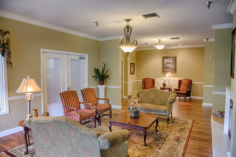 A cozy senior living facility common area with two green patterned sofas, two striped armchairs, a wooden coffee table with a flower arrangement, a side table with a lamp, and two additional armchairs with a small table and lamp in the background. The room has wooden flooring, beige walls with white trim, and ceiling light fixtures.