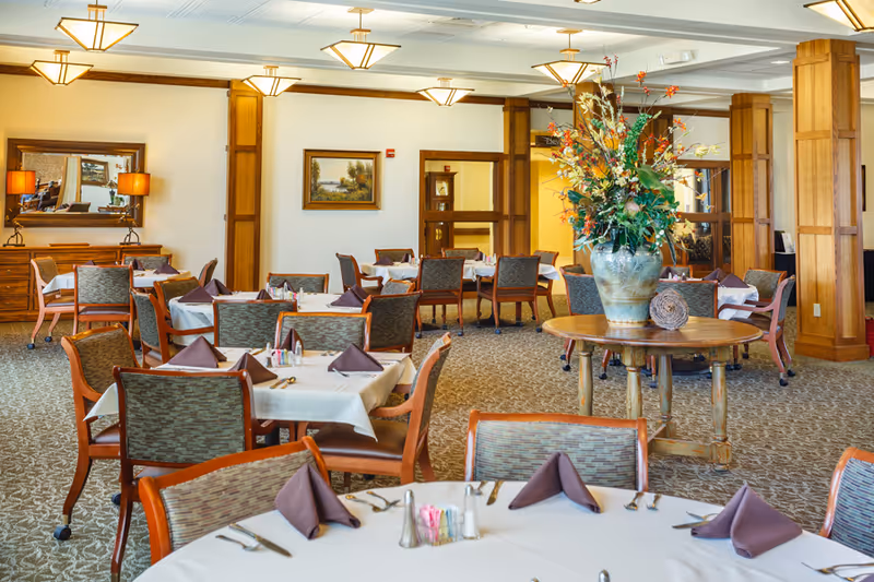 A spacious dining room in a senior living facility with multiple tables covered in white tablecloths and set with brown folded napkins, silverware, and condiments. The room features wooden chairs with patterned upholstery, a large floral arrangement on a round wooden table in the center, beige carpet, and warm lighting from ceiling fixtures. There are paintings and mirrors on the walls, and wooden columns add to the decor.