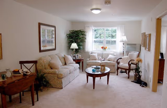A cozy living room with two beige patterned sofas, a wooden coffee table with decorative items, a wooden side table with a lamp and picture frames, a wooden chair, a potted plant, and a window with white curtains letting in natural light.