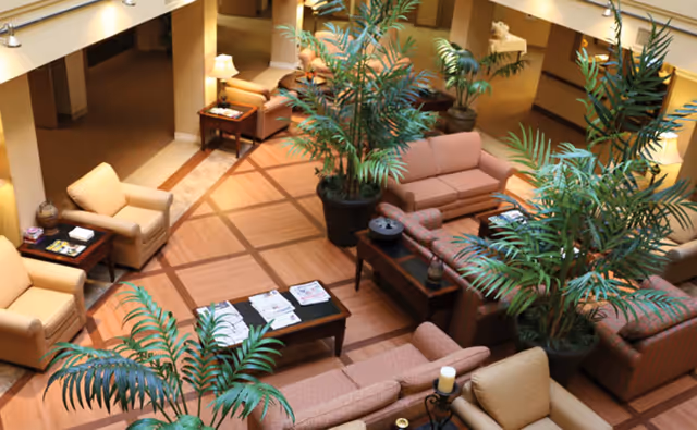A spacious and well-lit living room area with multiple beige and light brown sofas and armchairs arranged around wooden coffee tables. Several large potted plants add greenery to the space. The floor has a patterned wood design, and there are lamps providing warm lighting throughout the room.