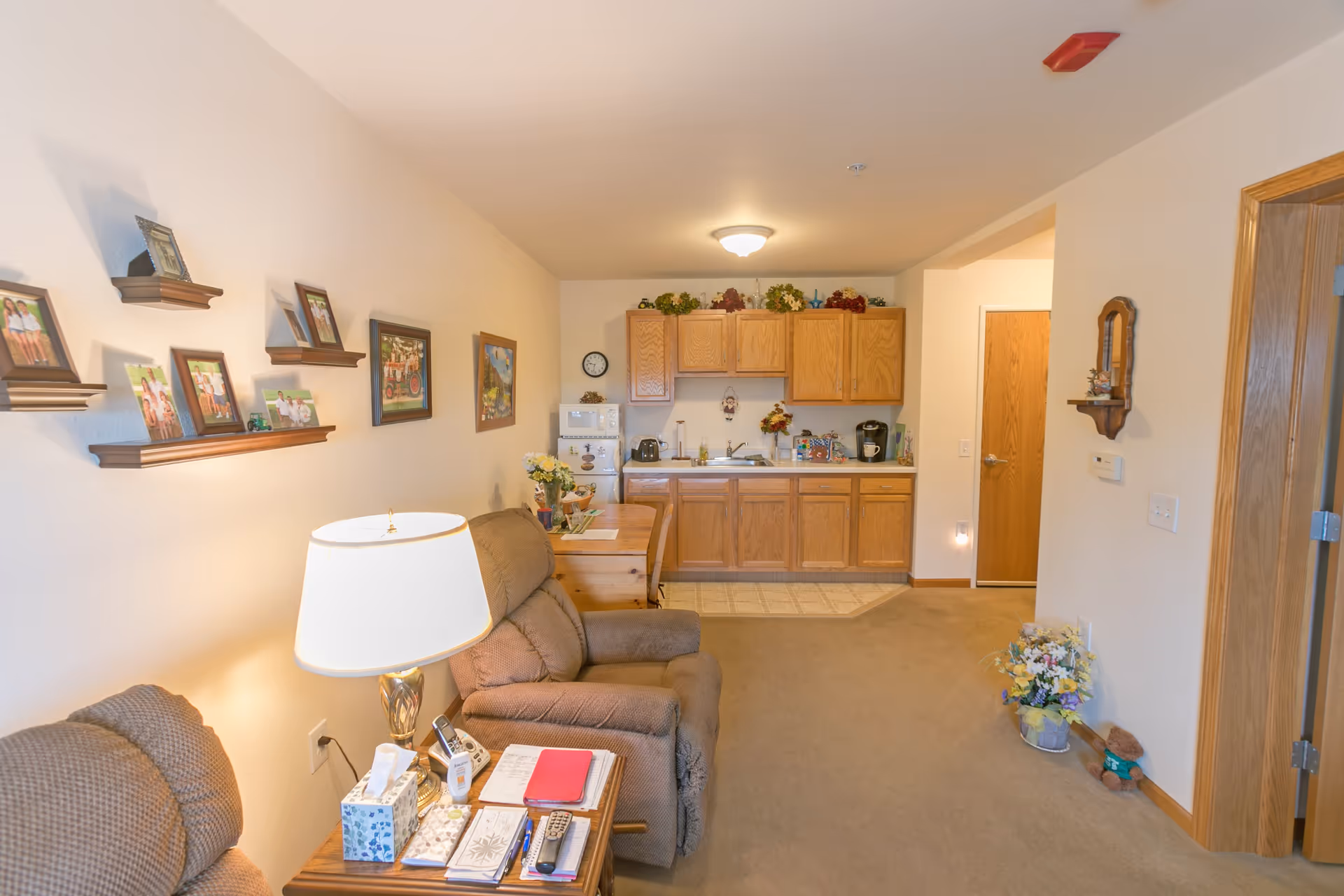 A cozy living area with two brown recliner chairs and a wooden side table holding a lamp, tissues, remote controls, and notebooks. On the wall above the chairs are several framed photos and paintings on small wooden shelves. In the background, there is a small kitchen area with wooden cabinets, a microwave, a toaster, a coffee maker, and decorative plants on top of the cabinets. The room has beige walls and carpet, with a wooden door and a small shelf with a mirror on the right side.