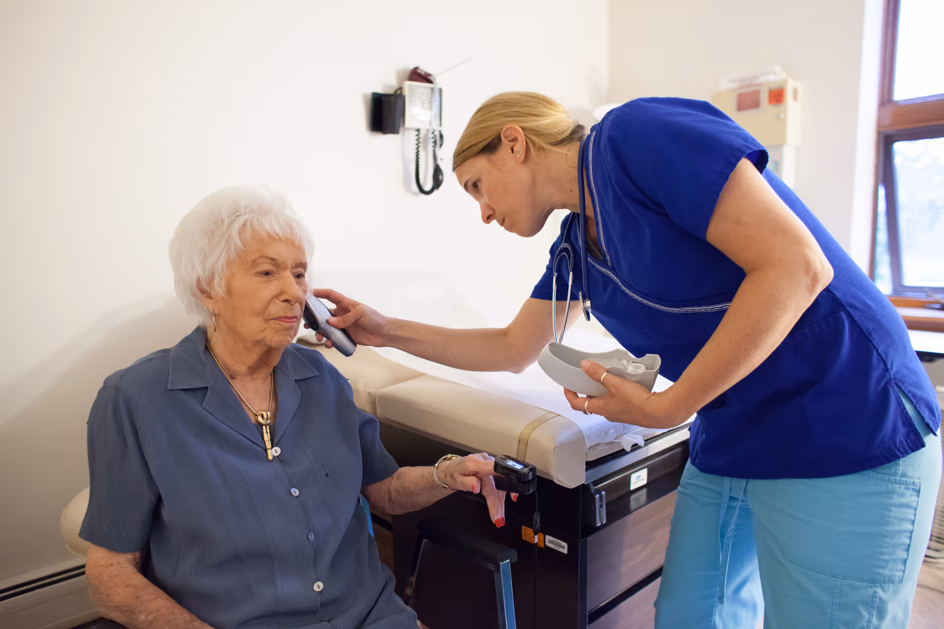 A healthcare professional in blue scrubs is examining an elderly woman with white hair using a medical device on her ear in a clinical room with an examination table and medical equipment.