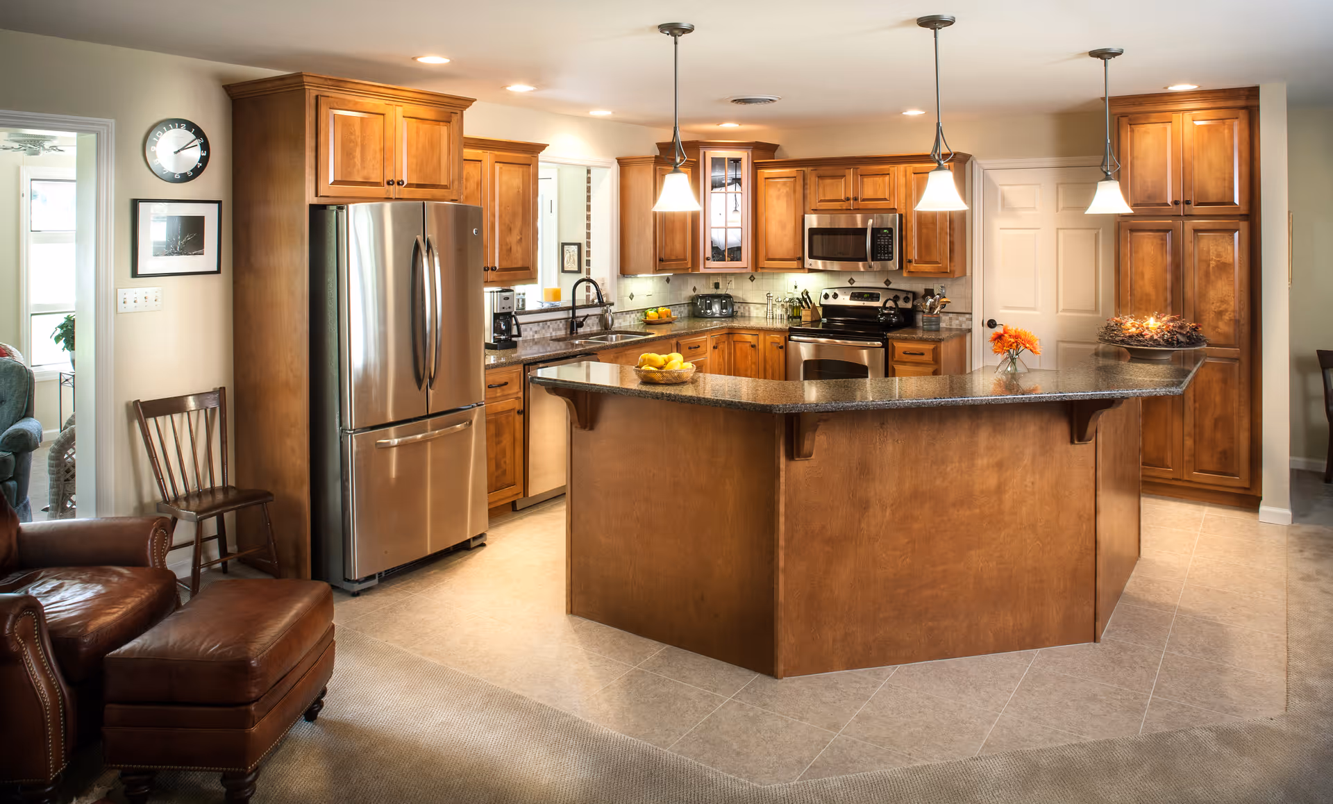 A spacious kitchen with wooden cabinets and a large granite countertop island. The kitchen features stainless steel appliances including a refrigerator, microwave, and stove. There are three pendant lights hanging above the island. To the left, there is a wooden chair and a leather armchair with an ottoman. The floor is tiled and the walls are painted in a light color.