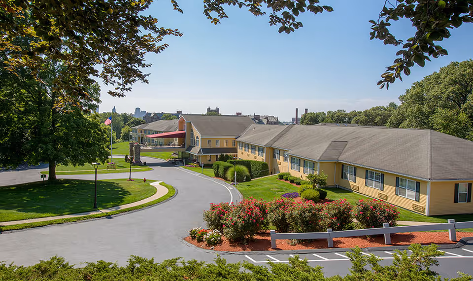 Exterior view of a senior living facility with yellow buildings, a curved driveway, well-maintained green lawns, flower beds, and trees under a clear blue sky.