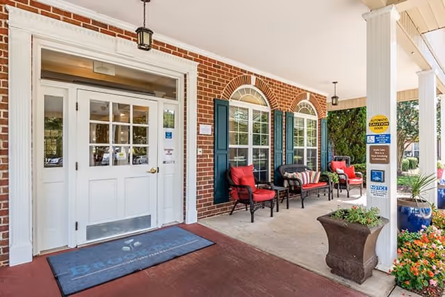 Covered entrance area of a building with white double doors, brick walls, and two large windows with green shutters. There are red cushioned chairs and a bench with striped pillows arranged on the porch, along with potted plants and a blue mat on the ground.
