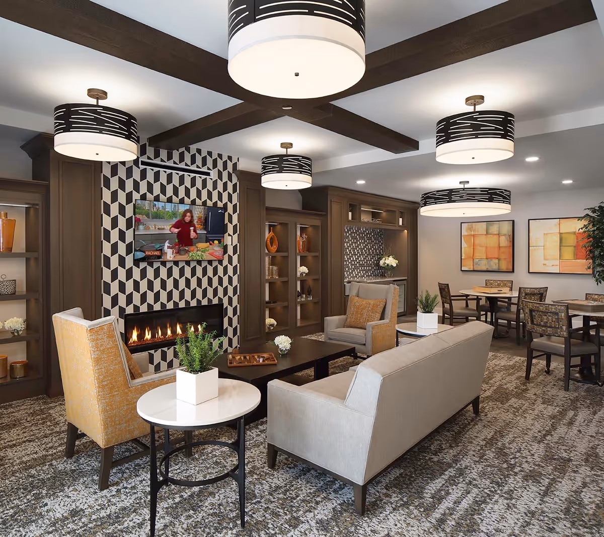 A cozy senior living common area with modern decor featuring a fireplace with a geometric patterned surround, a wall-mounted TV, beige and yellow upholstered chairs, a beige sofa, round and rectangular tables with small potted plants, built-in wooden shelves with decorative items, and abstract artwork on the walls. The ceiling has exposed wooden beams and multiple stylish pendant lights.