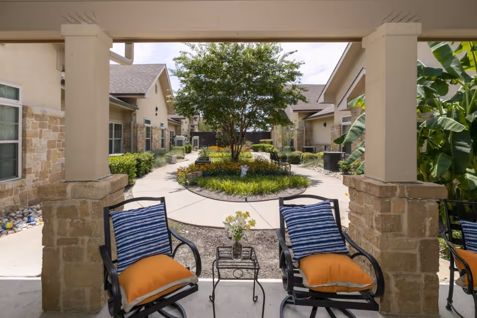 View from a covered patio area with two cushioned chairs and a small table with flowers, overlooking a landscaped courtyard with a tree, bushes, and a paved walkway surrounded by single-story buildings.