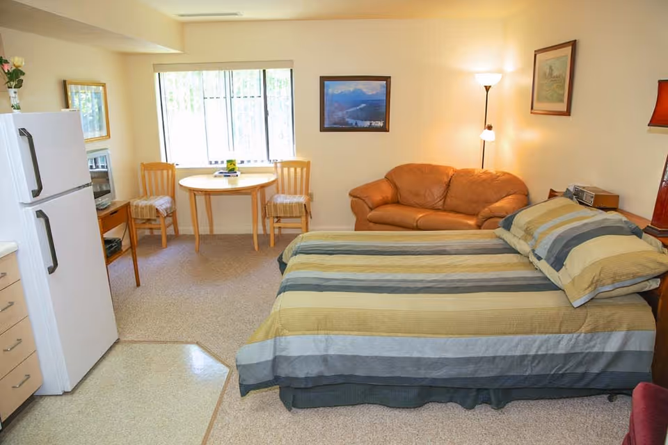 A cozy senior living studio apartment featuring a bed with striped bedding, a small round wooden table with two chairs near a window, a brown leather couch, a floor lamp, framed artwork on the walls, a white refrigerator, and a small TV on a wooden stand.