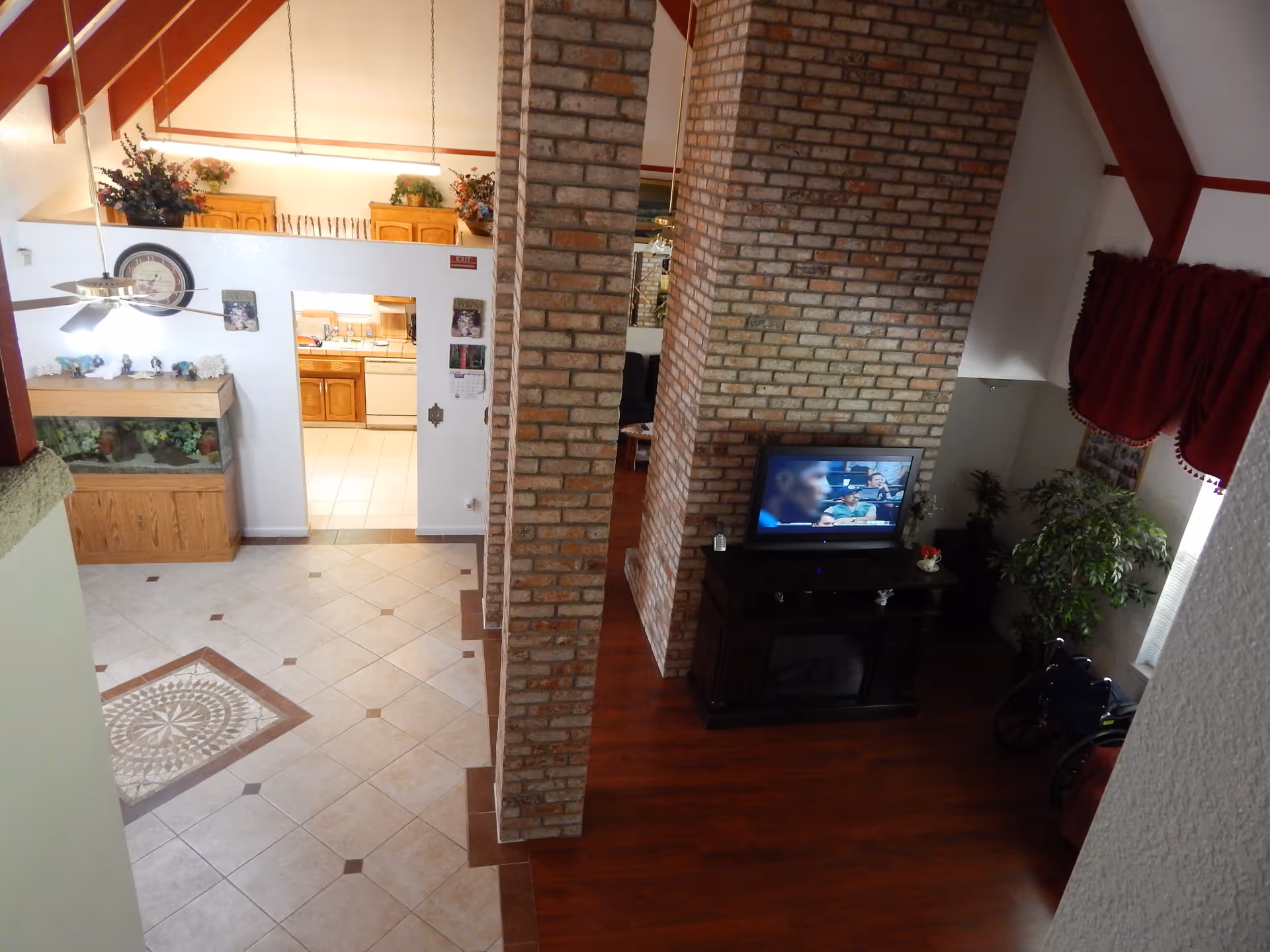 Interior view of a senior living facility showing a tiled floor area with a decorative tile pattern, a brick column dividing the space, a kitchen with wooden cabinets in the background, and a living area with a TV on a stand, a plant, and a wheelchair near a window with red curtains.