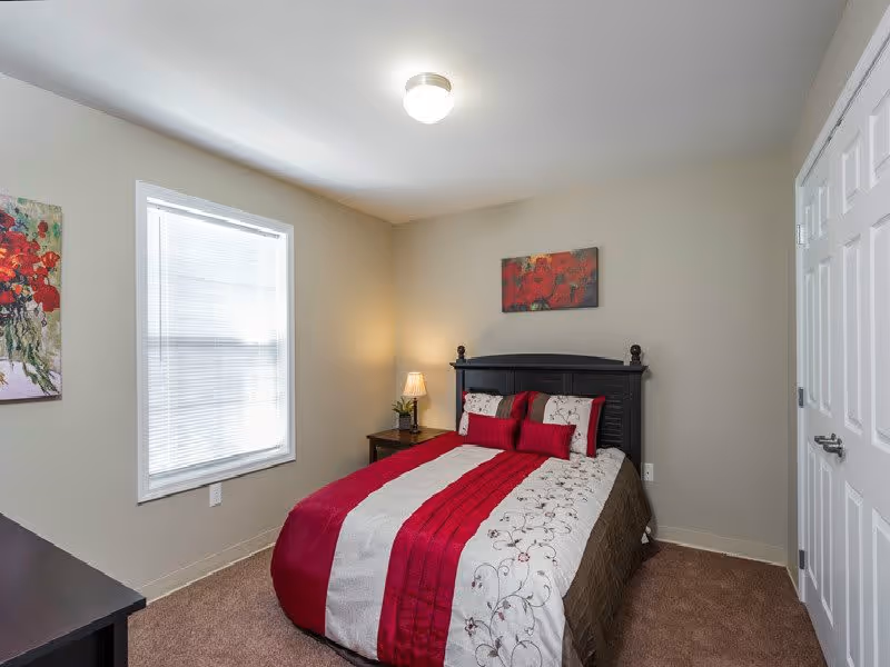 A bedroom with a bed featuring a red, white, and brown patterned comforter, two matching pillows, and a black headboard. There is a nightstand with a lamp and a small plant next to the bed. A window with blinds is on the left wall, and a colorful floral painting hangs above the bed. The walls are painted light beige, and the carpet is brown.