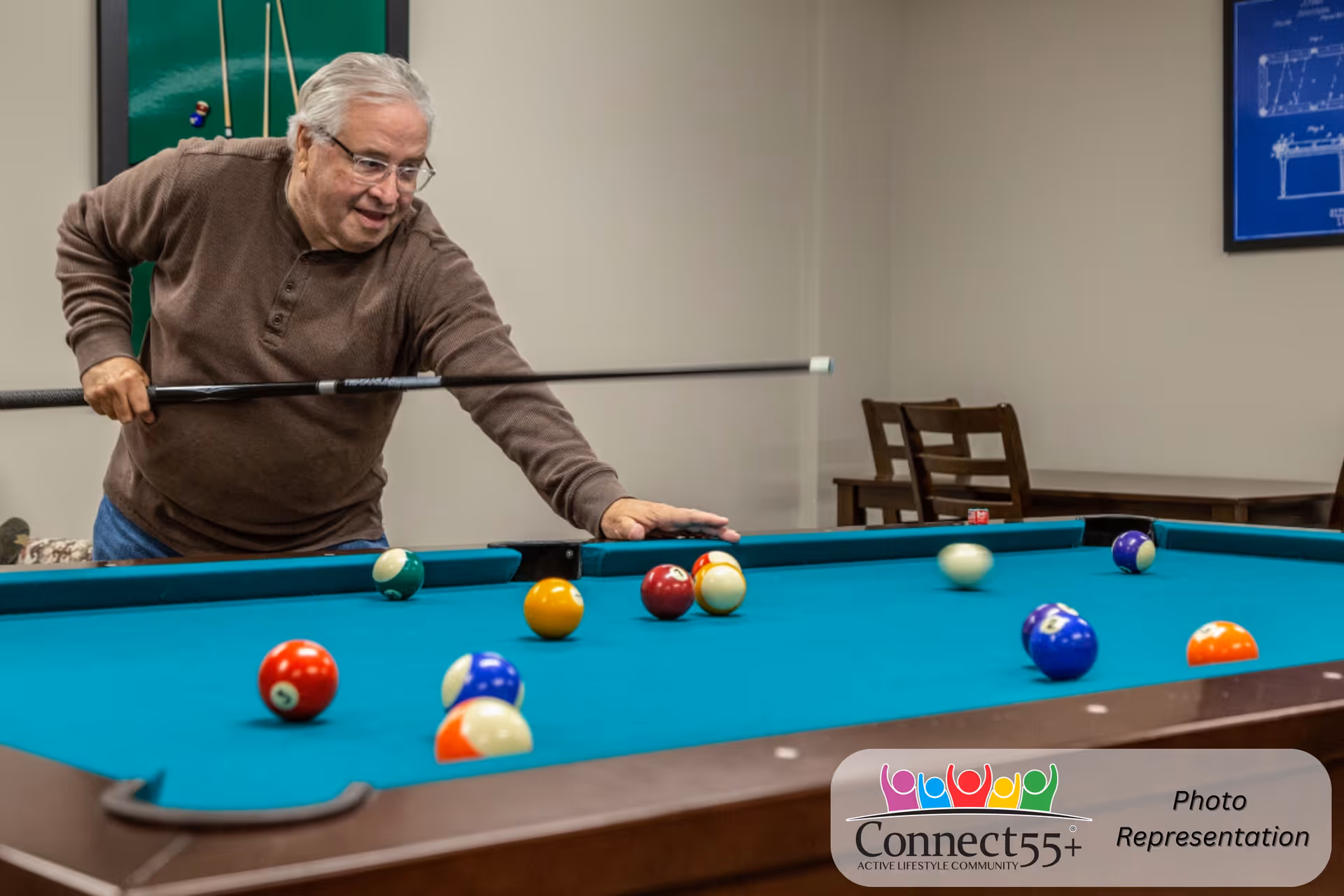An elderly man playing pool indoors, aiming with a cue stick at a billiard table with several colorful balls scattered on the green felt surface. In the background, there are chairs and framed pictures on the wall.