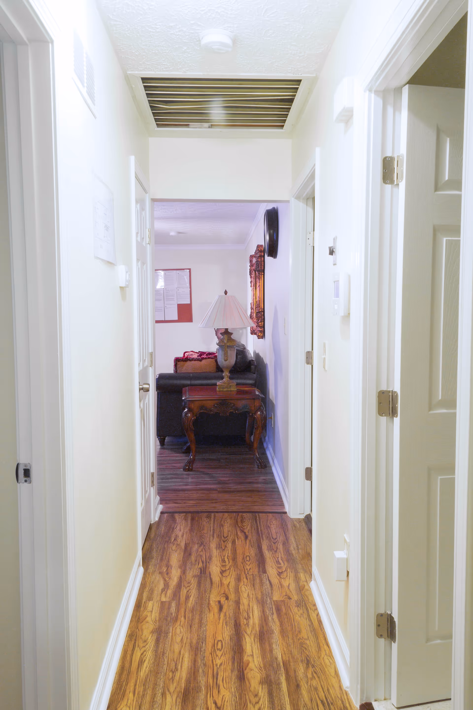 View down a hallway with wooden flooring leading to a living room area. The hallway has white walls and doors on both sides. At the end of the hallway, there is a wooden side table with a decorative lamp on it, and a couch with cushions is partially visible in the living room.