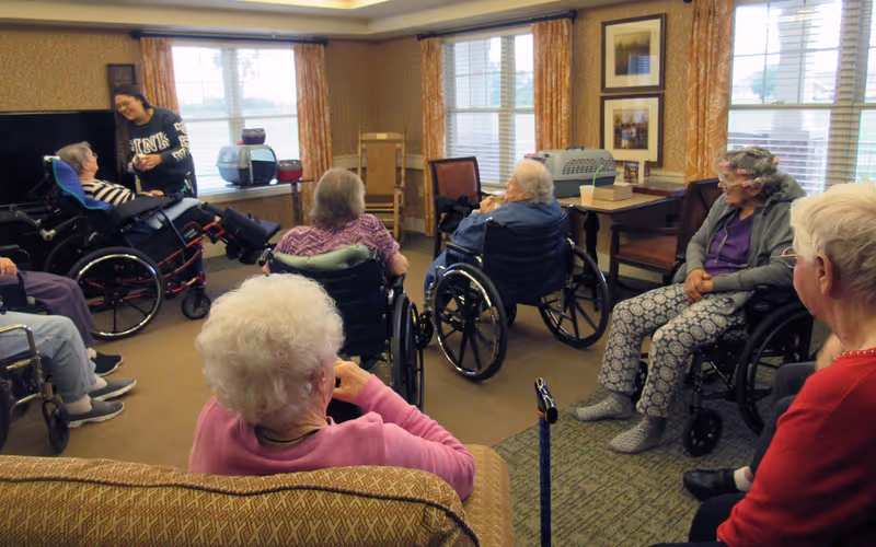 A group of elderly individuals, most in wheelchairs, gathered in a well-lit common room with large windows and patterned curtains. A younger woman is interacting with one of the seated elderly women. The room has comfortable seating, framed pictures on the walls, and a television.