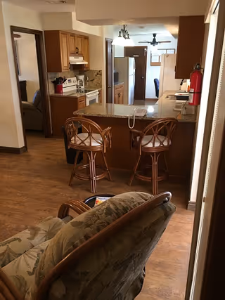 Open-plan assisted living interior with a kitchen island and two bar stools, stove and refrigerator in the background, and a cushioned chair in the foreground.