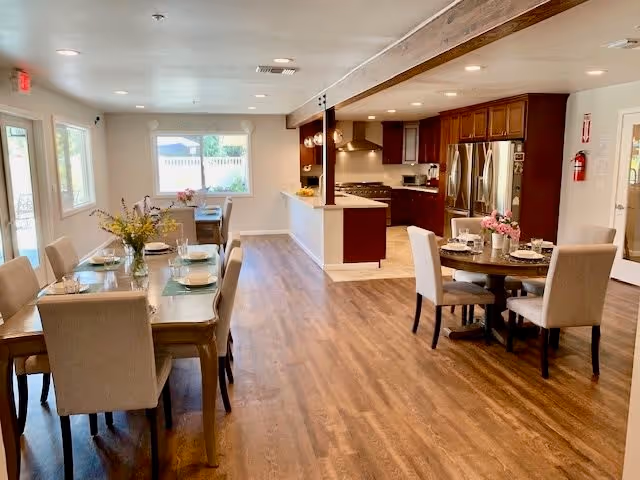 A spacious dining and kitchen area in a senior living facility with wooden flooring. The dining area has two tables set with plates, glasses, and floral centerpieces. The kitchen features dark wood cabinets, stainless steel appliances including a refrigerator and stove, and a countertop with bar seating. Large windows allow natural light to brighten the room.