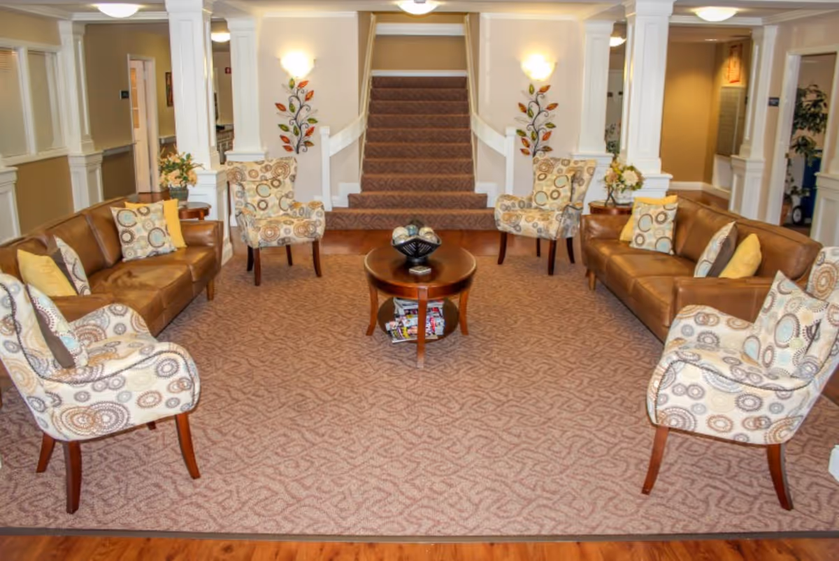 A cozy living room area with two brown leather sofas and four patterned armchairs arranged around a round wooden coffee table. The room features beige walls with white trim, decorative wall sconces, and a carpeted staircase in the background. There are decorative pillows on the sofas and a floral arrangement on a side table.