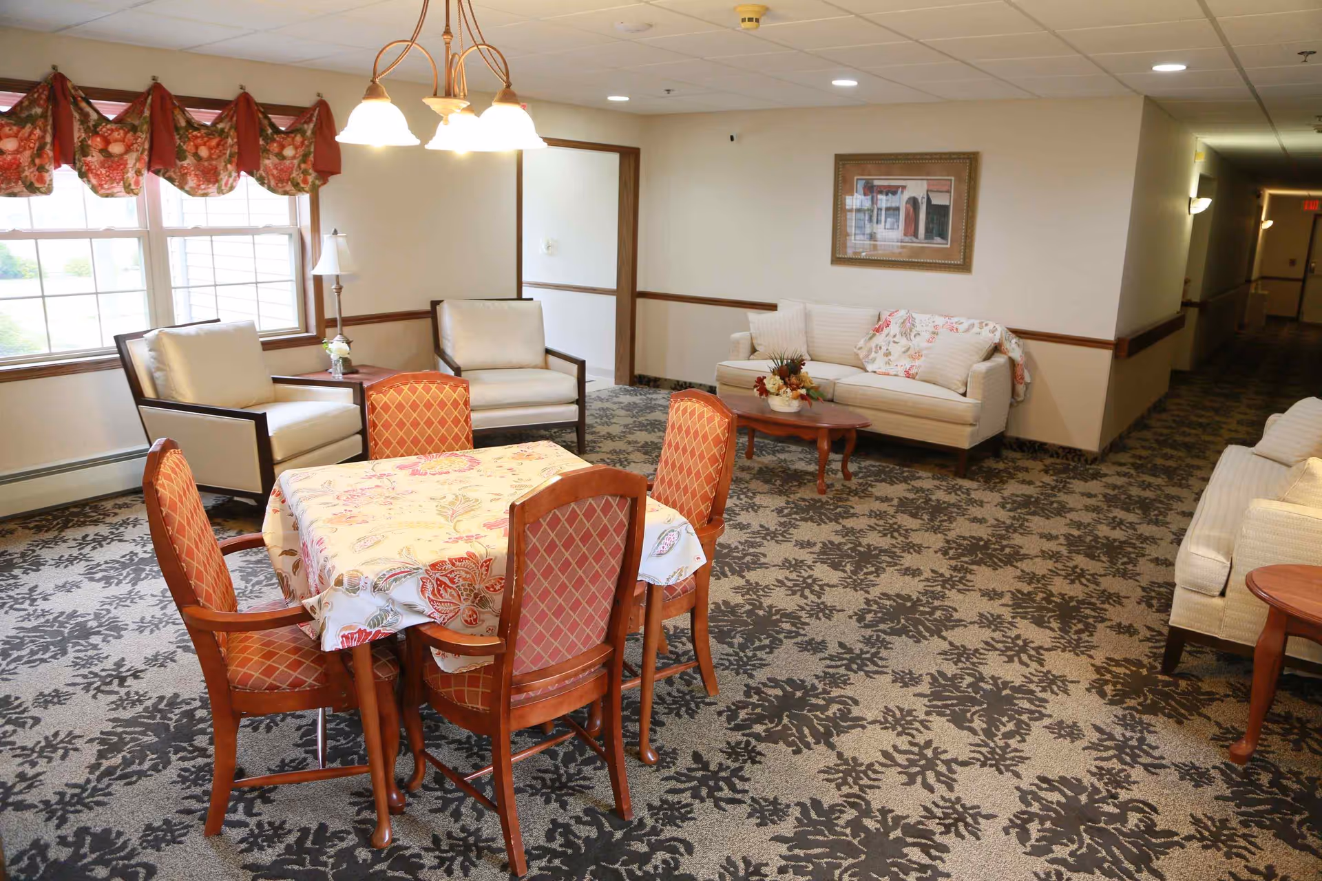 A bright communal sitting area with a table and four chairs, upholstered sofas and armchairs, floral-pattern carpet and a window with valance.